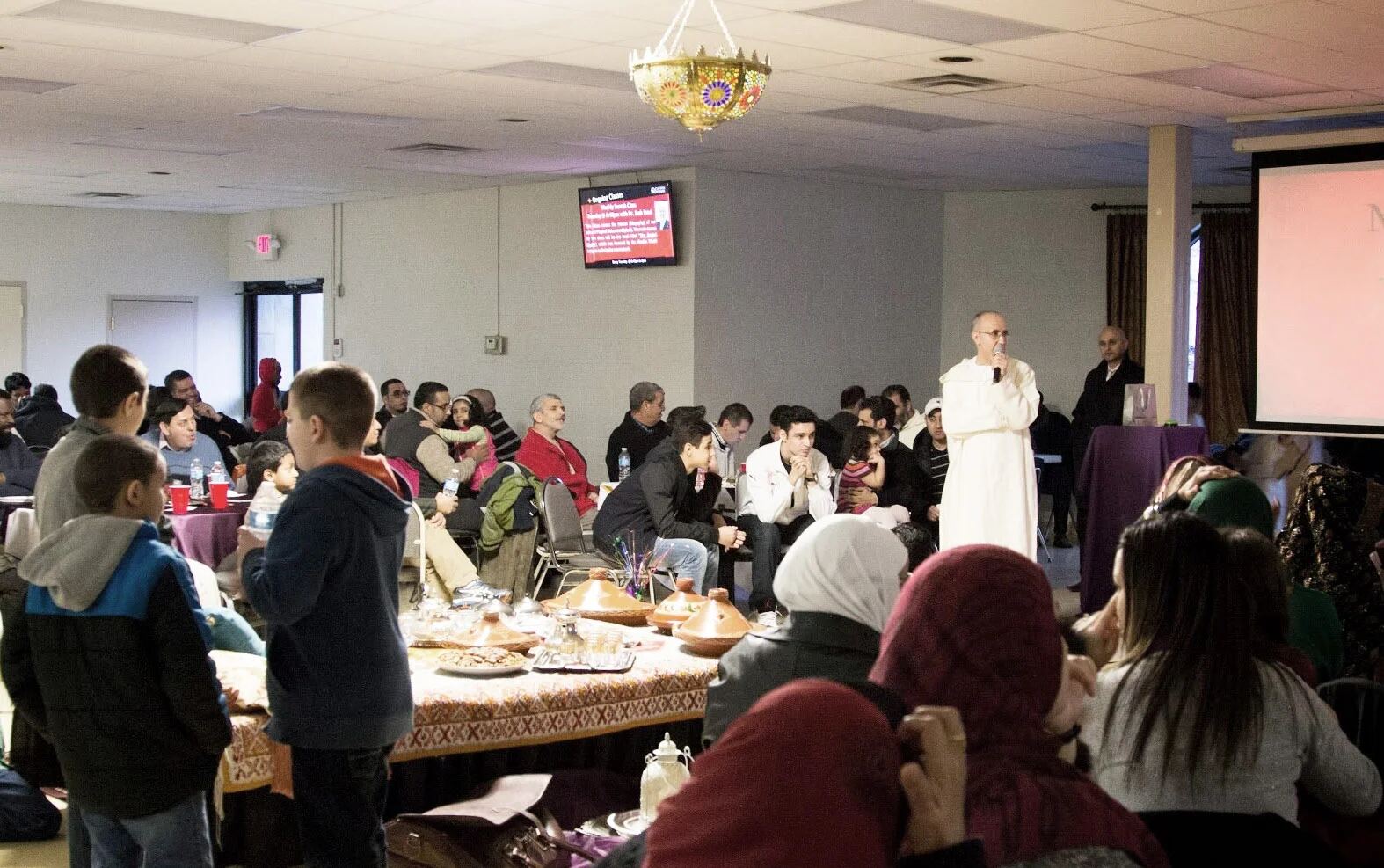 A full hall at Clifton Mosque: families at long tables set with tagines while a speaker in a white robe addresses the room